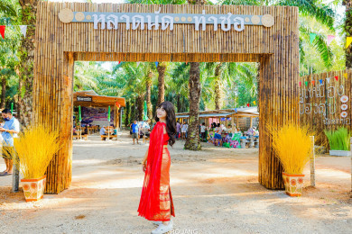 ผู้หญิงสวมชุดไทยสีแดงยินโพสถ่ายรูป หน้าซุ้มไม้ไผ่ที่มีป้าย “หลาดนัด ท่าซอ” ที่เที่ยวแห่งใหม่ของจังหวัดพังงา